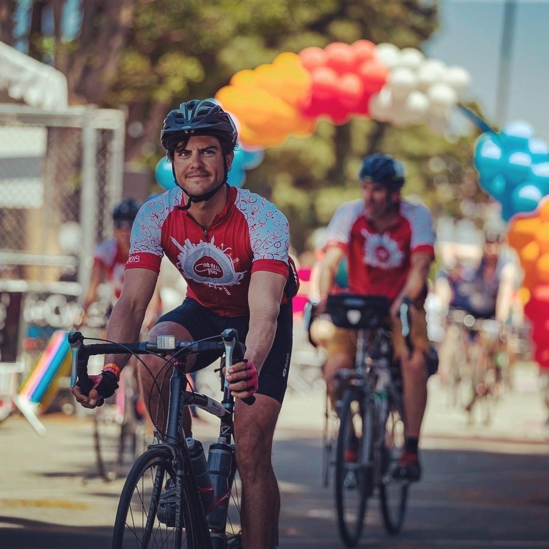 Geoff riding toward the camera in a red jersey, with balloons and other riders behind.