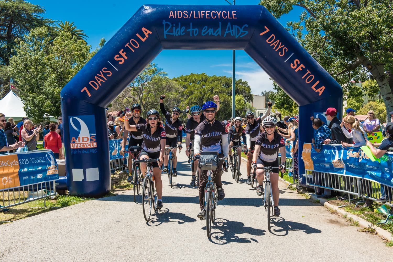 The Cabrio Taxi team, fists raised, riding under the AIDS/LifeCycle arch as crowds cheer.