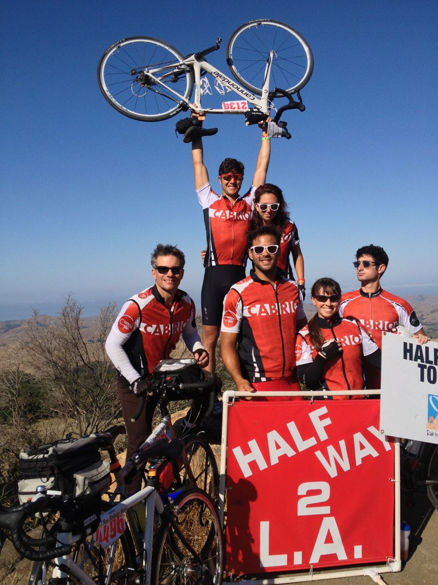 Six riders in red Cabrio jerseys at a hilltop sign reading Halfway 2 L.A., one holding a bike overhead.