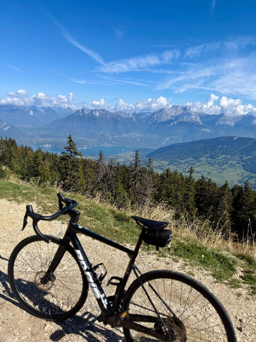 A road bike on a gravel track high above Lake Annecy, with forest below and mountains across the lake.