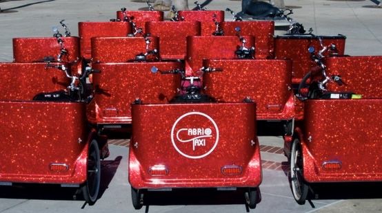 A dozen sparkling red pedicabs parked in rows, one showing the Cabrio Taxi logo.