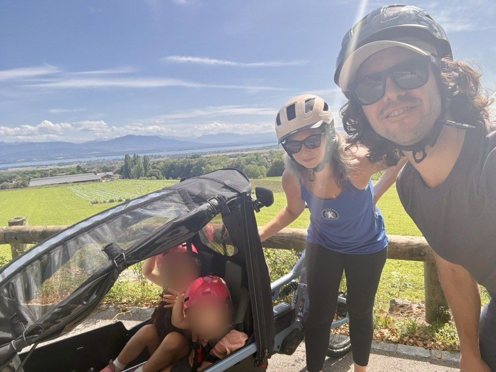 Geoff, his wife and their two children in a cargo bike, stopped above vineyards and a lake with mountains behind.