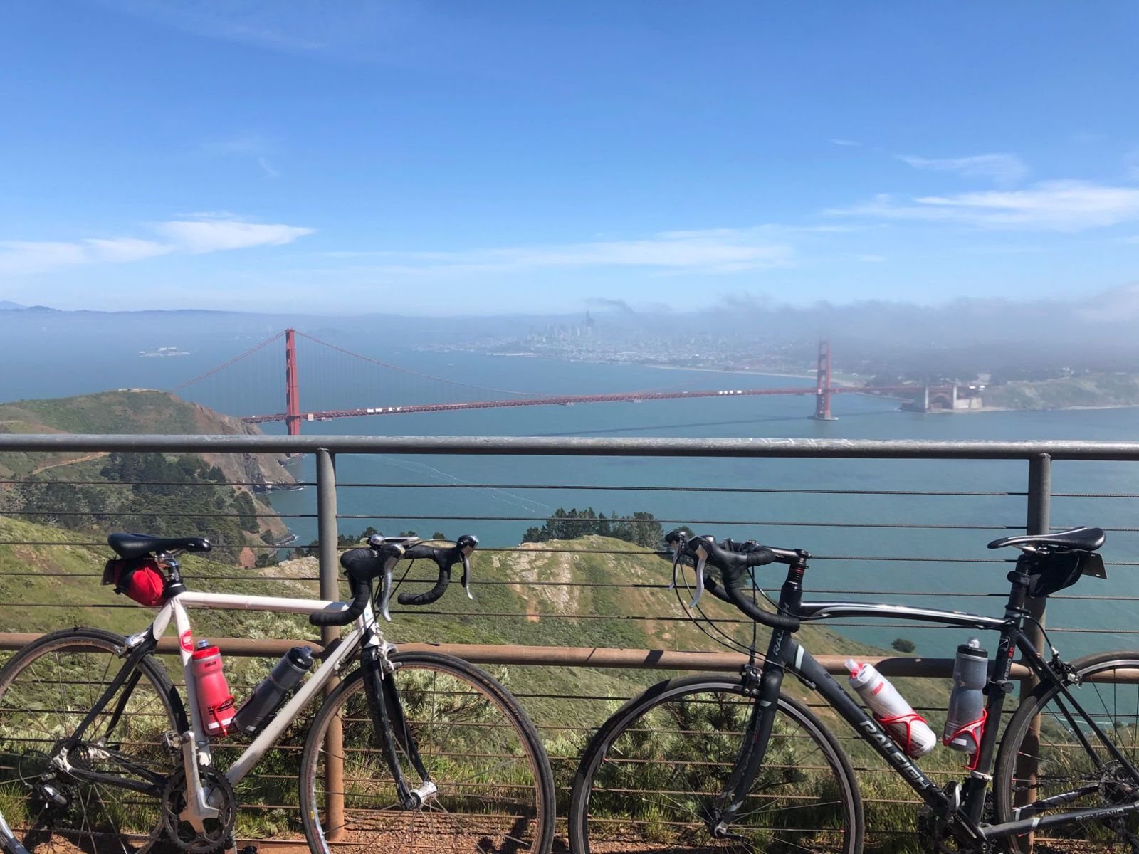 Two road bikes against a railing above the Golden Gate Bridge, with San Francisco in fog behind.