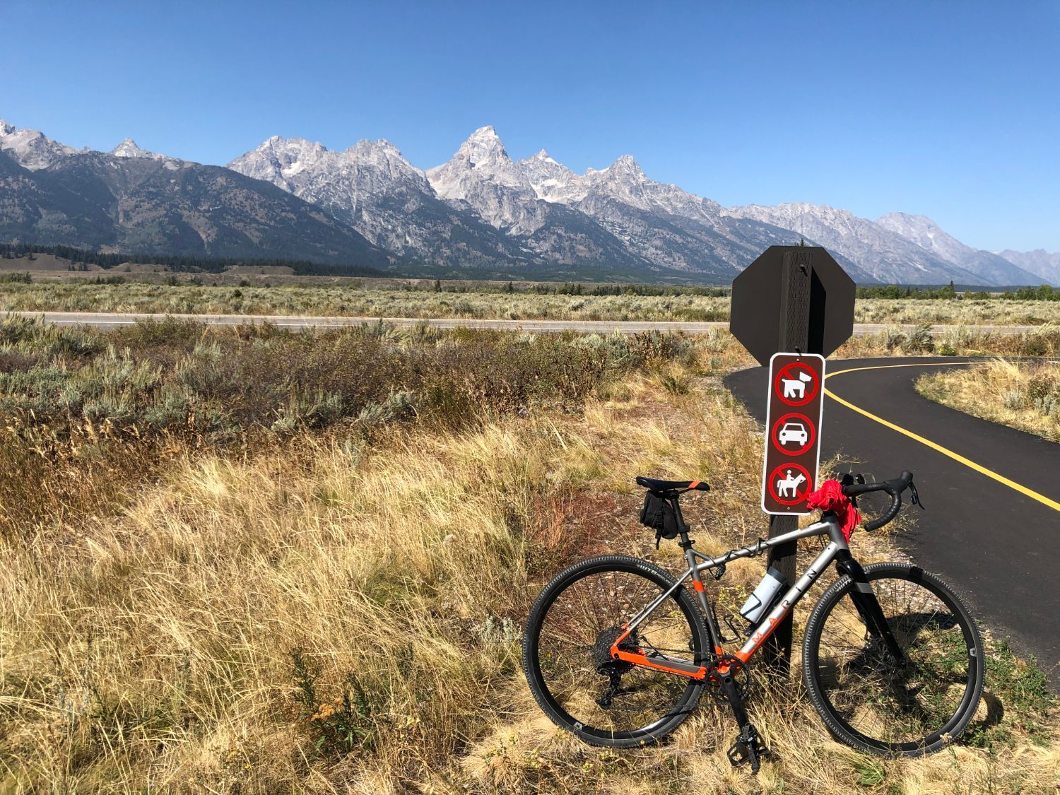 A gravel bike against a trail sign on a bike path, with the Teton range behind.