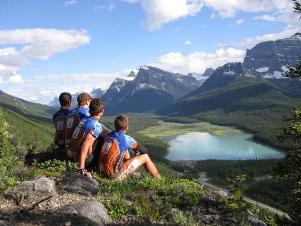 Four riders in team jerseys sit on a ridge looking over a turquoise lake and mountains.