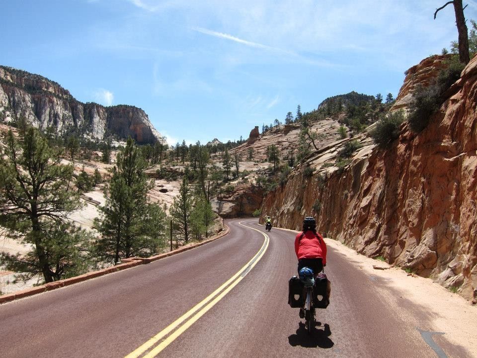 A loaded touring bike on a red road winding between sandstone cliffs.